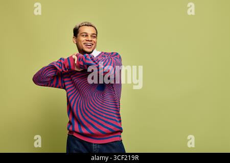 Ein stylischer junger Mann lächelt selbstbewusst in einem gestreiften Sweatshirt auf grünem Hintergrund. Stockfoto