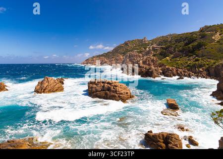 Türkisfarbene Wellen prallen an der zerklüfteten Küste von Costa Paradiso, Sardinien, unter einem leuchtend blauen Sommerhimmel auf massive rötliche Felsen. Stockfoto