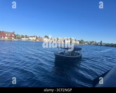 Ein kleines überdachtes Boot fährt an einem sonnigen Tag über blaues Wasser vor farbenfrohen Häusern am Wasser, mit klarem Himmel und fernen modernen Gebäuden in t Stockfoto