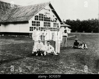 Dieses Gruppenporträt aus dem Jahr 1900-1920 zeigt Menschen auf einem Tennisplatz neben einem Tabak- oder Gärschuppen. Das Ambiente spiegelt soziale Aktivitäten während der Kolonialzeit in Indonesien wider. Stockfoto