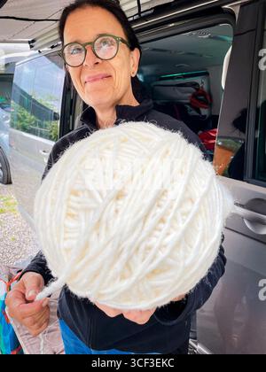 Eine dunkelhaarige Frau mit Brille hält einen weißen Wollball vor der Kamera vor einem Wohnmobil auf einem Campingplatz Stockfoto