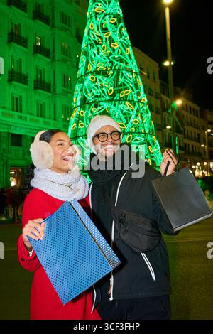 Fröhliches Paar, das Einkaufstaschen hält und lächelt, während es bei der weihnachtsfeier in der Stadt in der Nähe des beleuchteten weihnachtsbaums steht Stockfoto