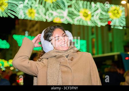 Glückliche junge Frau passt ihre Ohrenschützer an und genießt den weihnachtsgeist unter dekorativen Lichtern Stockfoto