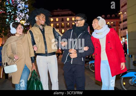 Vier Freunde gehen Arm in Arm und lachen nachts gemeinsam auf einer Stadtstraße und genießen die Wintersaison Stockfoto