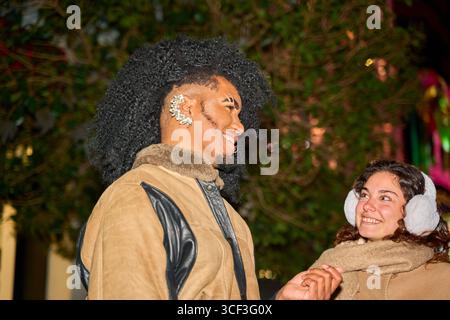 Junger Mann mit extravaganter Frisur, der Hände hält mit einer Frau, die Ohrenschützer in einer städtischen Umgebung trägt Stockfoto