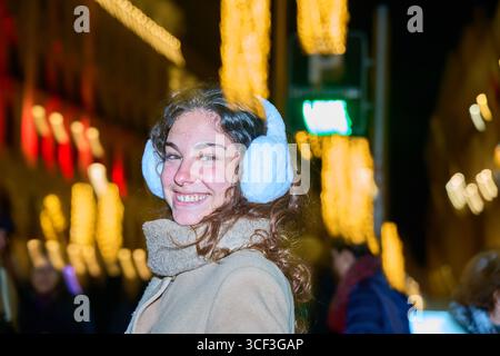 Fröhliche junge Frau lächelt, während sie Ohrenschützer trägt und die festliche Atmosphäre eines geschäftigen weihnachtsmarktes unter funkelnden Lichtern genießt Stockfoto