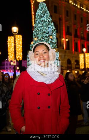 Junge Frau mit Ohrenschützern und rotem Mantel, die nachts auf einem weihnachtlich geschmückten Stadtplatz lächelt Stockfoto