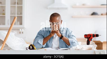 Probleme mit der Möbelmontage. Verstörter schwarzer Mann, der im Kücheninnenraum mit Werkzeugen sitzt und Anweisungen ansieht Stockfoto