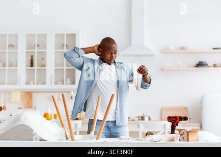 Verwirrt schwarzen Mann hält Paket mit Details, Montage Tisch mit Anweisungen, in der Küche Innenraum, kopieren Raum. Stockfoto
