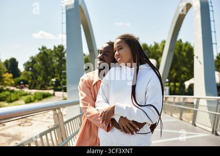 Ein liebevolles Paar umarmt sich an einem schönen sonnigen Tag, während er auf einer Brücke im Park spaziert. Stockfoto
