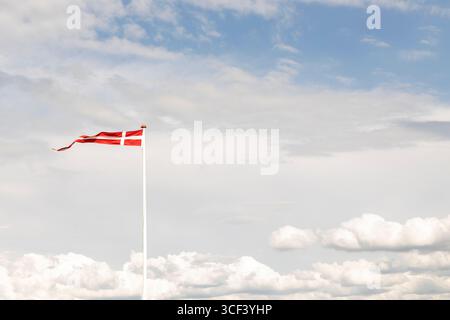 Dänische Flagge im Sonnenlicht auf einem Fahnenmast im Vordergrund winkend im Wind vor einem schönen blauen Himmel mit weißen Wolken, Insel Römo, Dänemark Stockfoto
