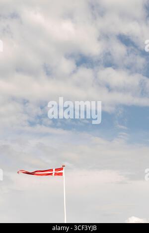 Dänische Flagge im Sonnenlicht auf einem Fahnenmast im Vordergrund winkend im Wind vor einem schönen blauen Himmel mit weißen Wolken, Insel Römo, Dänemark Stockfoto