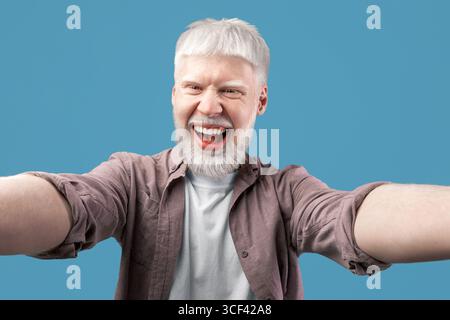 Fröhlicher Albino-Typ mit ungewöhnlichem Auftritt, der Selfie auf türkisfarbenem Studiohintergrund macht, die Kamera anlächelt und Spaß hat Stockfoto