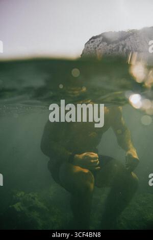 Ein Mann sitzt im Wasser mit den Händen auf den Knien. Das Wasser ist grün und trüb Stockfoto
