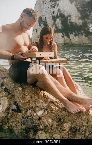 Ein Mann und eine Frau sitzen auf einem Felsen am Wasser und öffnen Pizzaschachteln. Die Szene ist entspannt und zwanglos, während das Paar seine Mahlzeit in einem wunderschönen n Stockfoto