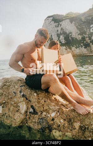 Ein Mann und eine Frau sitzen auf einem Felsen am Wasser und öffnen einen Karton. Die Szene ist entspannt und zwanglos, während das Paar ihre gemeinsame Zeit genießt Stockfoto