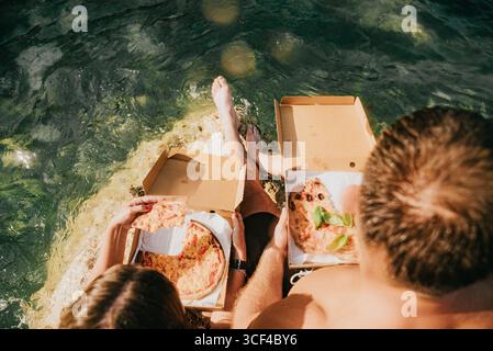 Ein Mann und eine Frau sitzen im Wasser mit zwei Kisten Pizza. Die Szene ist unbeschwert und lustig, während das Paar seine Pizza genießt, während es umgeben ist Stockfoto