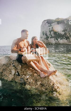 Ein Mann und eine Frau sitzen auf einem Felsen am Wasser und essen Pizza. Die Szene ist entspannt und zwanglos, während das Paar seine Mahlzeit in einer wunderschönen Natur genießt Stockfoto