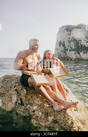 Ein Mann und eine Frau sitzen auf einem Felsen am Wasser und essen Pizza. Die Szene ist entspannt und zwanglos, während das Paar seine Mahlzeit in einer friedlichen Umgebung genießt Stockfoto