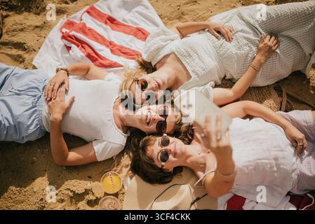 Drei Frauen liegen am Strand, eine von ihnen macht ein Selfie mit ihrem Handy. Die Szene ist entspannt und sorglos, während die Frauen ihre t genießen Stockfoto