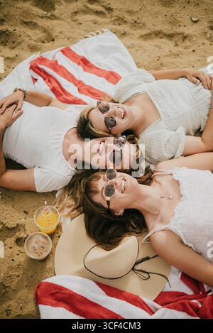 Drei Frauen liegen auf einem Strandtuch, lächeln und tragen eine Sonnenbrille. Es gibt Becher und einen Hut in der Nähe. Die Szene ist entspannt und sorglos Stockfoto