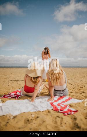 Drei Frauen sitzen am Strand, eine von ihnen fotografiert die anderen zwei. Die Szene ist entspannt und sorglos, während die Frauen ihre Zeit genießen Stockfoto