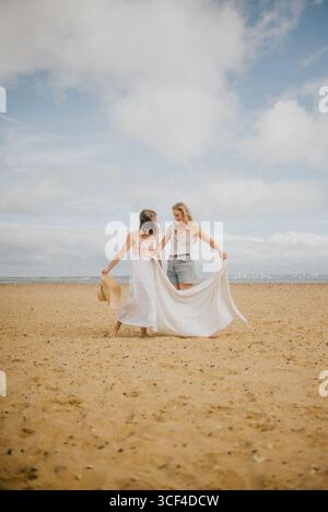 Zwei Frauen laufen am Strand, eine mit weißem Kleid. Die Szene ist unbeschwert und sorglos, während die beiden Frauen ihre Zeit genießen Stockfoto