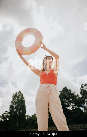Eine Frau hält einen pinkfarbenen und gelben aufblasbaren Ring. Sie trägt ein rotes Oberteil und eine weiße Hose. Die Szene spielt in einem Park mit Bäumen im Hinterland Stockfoto