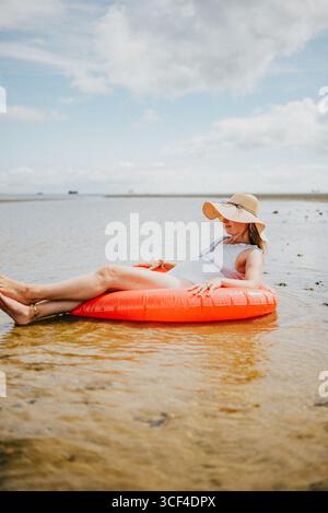 Eine Frau schwimmt auf einem roten Schlauchboot im Ozean. Das Wasser ist ruhig und der Himmel ist klar. Die Frau trägt einen Strohhut und einen weißen Tank Stockfoto