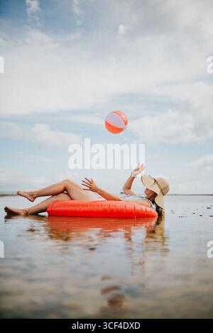 Eine Frau schwimmt auf einem roten Schlauchboot im Ozean. Sie hält einen orangen Strandball Stockfoto