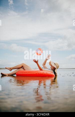 Eine Frau schwimmt in einem roten Schlauchboot mit einem Strandball in der Luft. Die Szene ist entspannt und sorglos, die Frau genießt ihre Zeit im Stockfoto