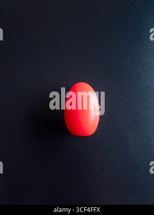 Minimalistisches Stillleben mit einer leuchtenden neonroten Tomate, die aufrecht vor einem dunklen, strukturierten Hintergrund steht und einen starken Kontrast erzeugt Stockfoto