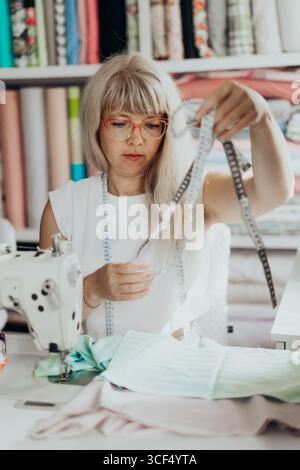 Die reife Frau steht in einem hellen Nähstudio mit Klebeband um den Hals, umgeben von bunten Stoffrollen, die lächeln, während sie an einem handgefertigten Stück arbeitet Stockfoto