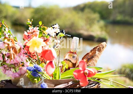 Fahrradtour mit Picknickkorb gefüllt mit Baguette, Wein und bunten Frühlingsblumen in einer sonnigen Uferlandschaft. Hintergrund für Urlaub und Freizeit. Stockfoto