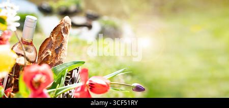 Fahrradtour mit Picknickkorb gefüllt mit Baguette, Wein und bunten Frühlingsblumen in einer sonnigen Uferlandschaft. Horizontaler Hintergrund für Urlaub und Freizeit. Leerzeichen für Text. Stockfoto