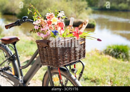 Fahrradtour mit Picknickkorb gefüllt mit Baguette, Wein und bunten Frühlingsblumen in einer sonnigen Uferlandschaft. Hintergrund für Urlaub und Freizeit. Stockfoto