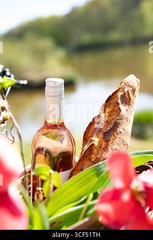 Fahrradtour mit Picknickkorb gefüllt mit Baguette, Wein und bunten Frühlingsblumen in einer sonnigen Uferlandschaft. Vertikaler Hintergrund für Urlaub und Freizeit. Leerzeichen für Text. Stockfoto