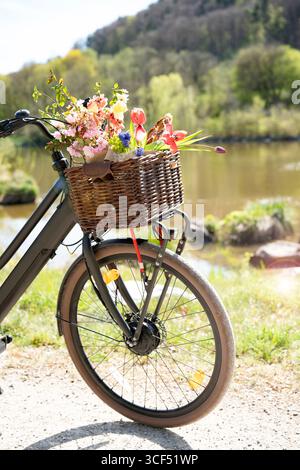 Ein Fahrrad mit bunten Blumen und mit Baguette gefülltem Korb steht auf einem Weg in einer idyllischen Frühlingslandschaft. Sonnige Fahrradtour im Hintergrund für umweltfreundlichen Tourismus, Freizeit und Lifestyle. Leerzeichen für Text. Stockfoto