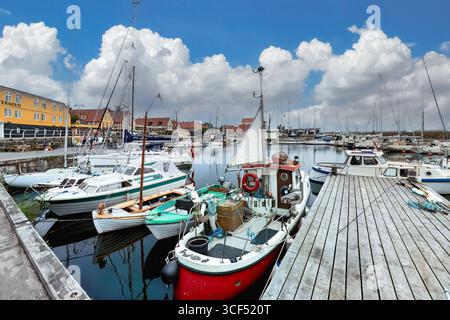 Farbenfrohe Boote legen entlang eines hölzernen Piers in einer lebhaften Hafenszene an, mit einem hellen Himmel und flauschigen Wolken, die eine ruhige Atmosphäre schaffen. Ronne, Bornh Stockfoto