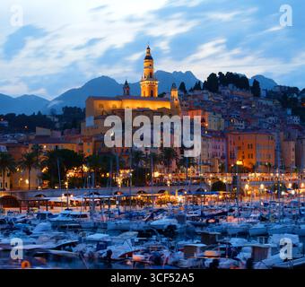 Panoramablick bei Nacht auf Menton, Frankreich, mit beleuchteter Altstadt, die sich auf dem Wasser spiegelt, im Vordergrund verankerten Yachten und den mediterranen Coas Stockfoto