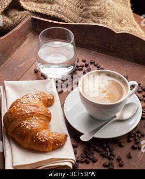 Cappuccino mit Croissant und ein Glas Wasser in die Schale mit dunkelbraunem Holz. Stockfoto