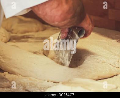 Baker arbeitet mit rohem Teig, teilt und formt ihn mit einem Metallschaber auf einer hölzernen Arbeitsfläche, bereitet das Backen vor und bereitet köstliches Brot zu Stockfoto