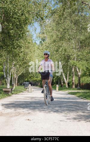 Hell beleuchtete Parklandschaft mit einer Person, die tagsüber auf einem Kiesweg radelt, umgeben von üppigen Bäumen und Grün. Stockfoto