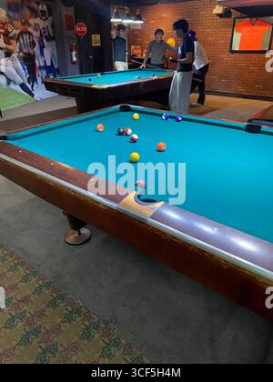 Eine Gruppe junger Freunde spielt Billard auf einem türkisfarbenen Filztisch in einer schwach beleuchteten Bar oder Erholungshalle, wobei ein Spiel im Vordergrund steht. Stockfoto