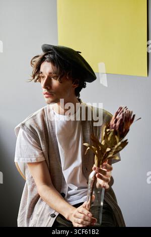 Vertikales Atelier-Porträt eines jungen Kaukasiers, der eine Baskenmütze trägt und eine Vase mit einer Blume darin hält, die wegblickt Stockfoto