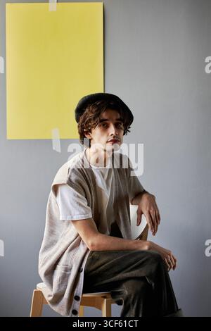 Studio-Porträt eines hübschen jungen kaukasischen Mannes in lässigem Outfit mit Baskenmütze, der vor grauem Wandhintergrund sitzt, mit gelbem Blatt Papier drauf und in die Kamera blickt Stockfoto