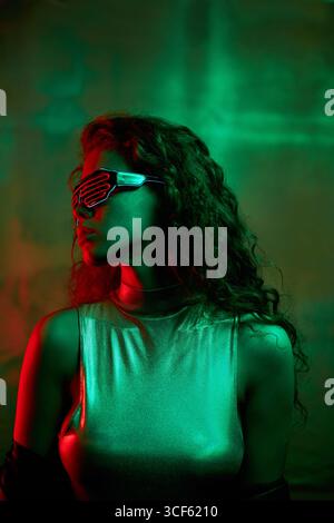 Porträt einer stilvollen jungen kaukasischen Frau mit langen lockigen Haaren, die eine futuristische Brille trägt, die in grünem und rotem Neonlicht steht und wegblickt Stockfoto