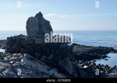 Basaltfelsen umrahmen ein verwittertes Steindenkmal an der Küste in der Nähe des Hafens Gonae, mit Blick über ruhiges Wasser und Gezeitenbecken in Richtung des offenen Meeres unter ihnen Stockfoto