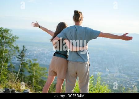 Junges Paar, das sich mit ausgestreckten Armen umarmt und während der Wanderung einen atemberaubenden Blick auf die Stadt vom Gipfel des Berges aus genießt. Reise-, Romantik- und Abenteuerkonzept. Stockfoto