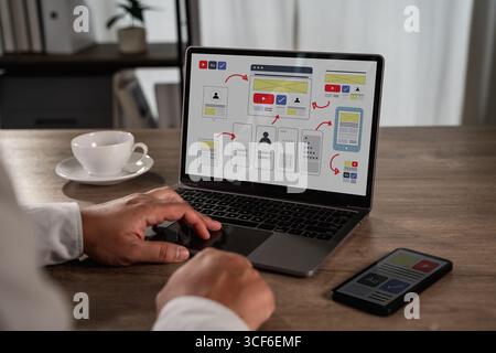 Das Konzept des UX-UI-Entwurfs zeigt einen Designer, der an einem Laptop arbeitet, mit Interfaces-Wireframes und User-Flow-Diagrammen, die die kreative Webentwicklung repräsentieren Stockfoto